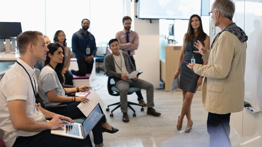 A few employees sitting near the front of a room taking notes and looking interested in a presentation by a colleague