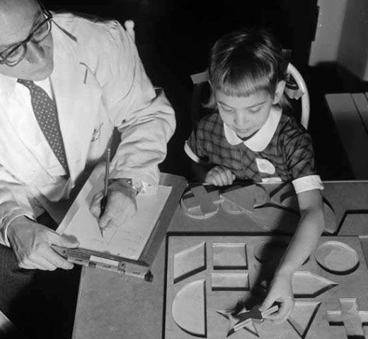 young girl matching shapes into a puzzle with a doctor watching and taking notes